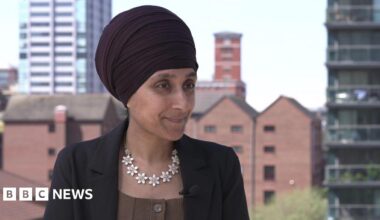 Sukhvinder Kaur is wearing a dark Sikh head covering or turban. He has a black jacket, with brown top and a striking necklace of white flowers.