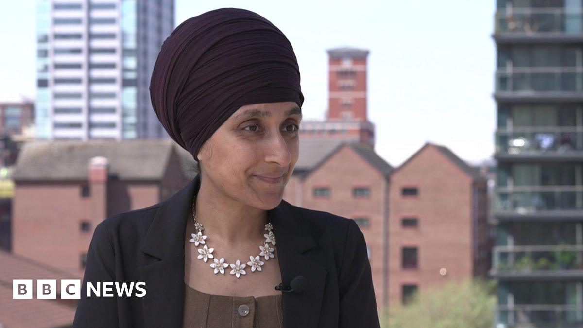 Sukhvinder Kaur is wearing a dark Sikh head covering or turban. He has a black jacket, with brown top and a striking necklace of white flowers.