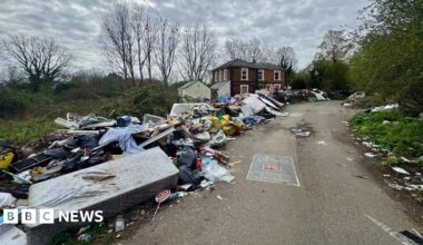 Fly-tipped rubbish piled up along a roadside leading to a red brick building and trees