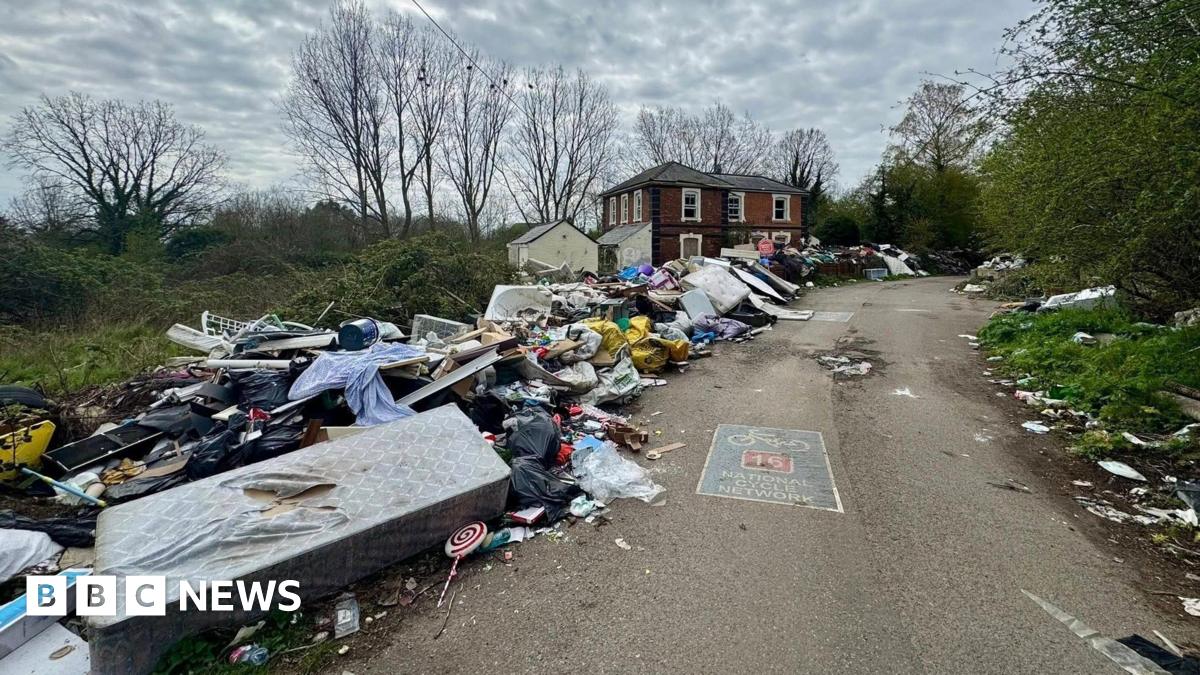 Fly-tipped rubbish piled up along a roadside leading to a red brick building and trees