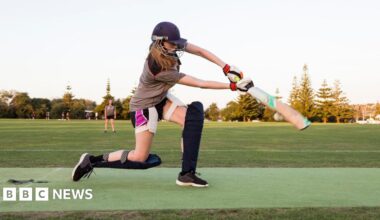 A girl bats wearing a helmet, shin pads and gloves. She is on a cricket pitch in the early evening