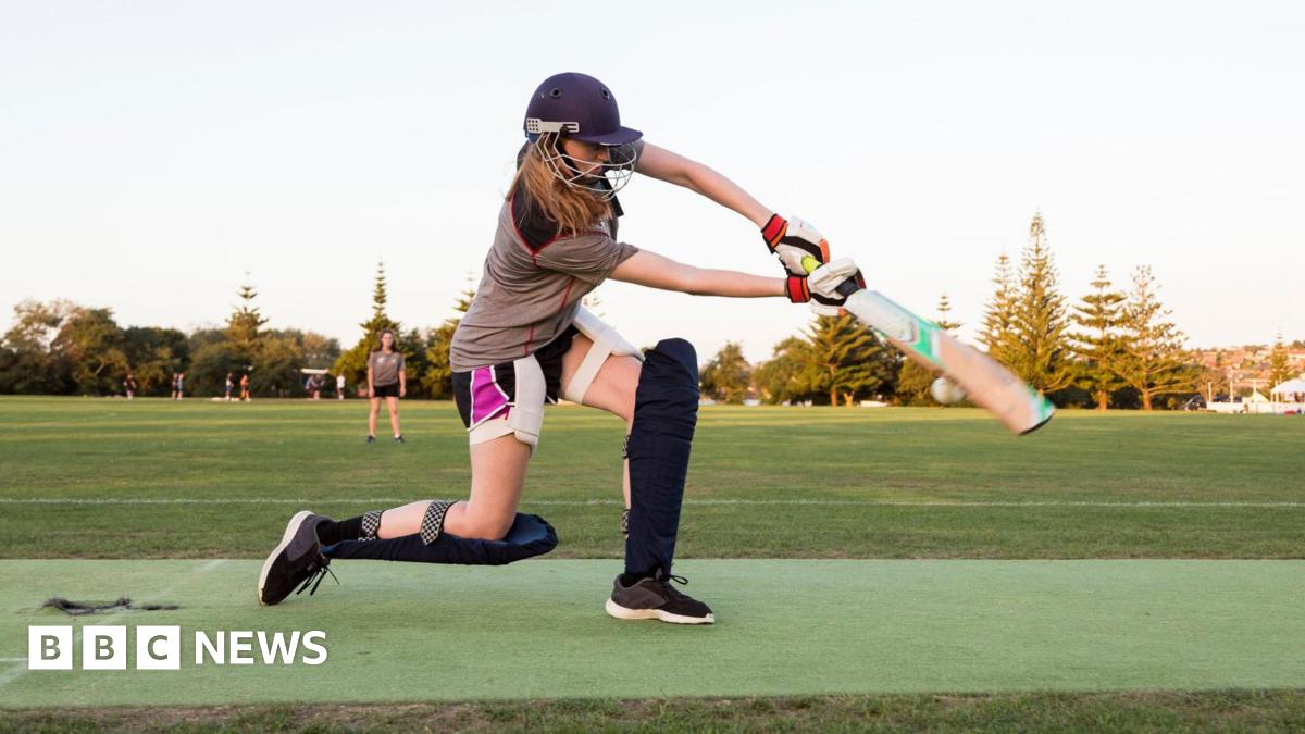 A girl bats wearing a helmet, shin pads and gloves. She is on a cricket pitch in the early evening
