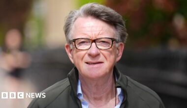 A man with grey hair and tortoiseshell glasses squints in the sunlight. He is wearing a khaki green fleece or coat over a light blue shirt