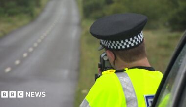 A uniformed police officer next to a single carriageway rural road with a speed gun.