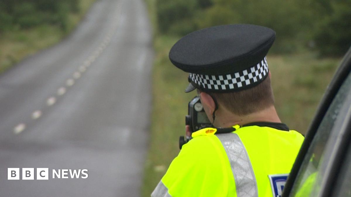 A uniformed police officer next to a single carriageway rural road with a speed gun.