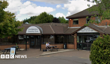 The entrance to Humberston Country Club. A man is sitting on a black bench outside of the entrance. They sky is blue with white clouds.