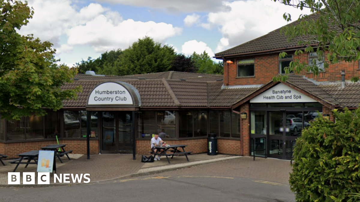 The entrance to Humberston Country Club. A man is sitting on a black bench outside of the entrance. They sky is blue with white clouds.