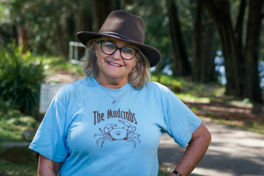 Ms Gibbs wearing a brown hat and a blue 'Mudcrabs' shirt stands next to a path in the shade.