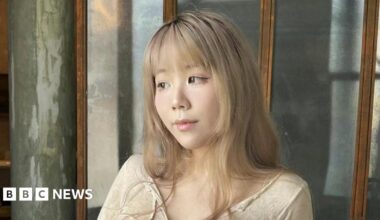 A woman with mid-length blonde hair and a fringe looks to her right, wearing a beige top and winged eyeliner.