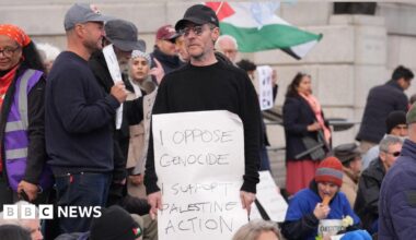 Massive Attack frontman Robert Del Naja standing at a protest at Tralfalgar Square holding a sign that reads: "I oppose genocide I support Palestine Action." He is wearing a black baseball cap, sunglasses and a black jumper