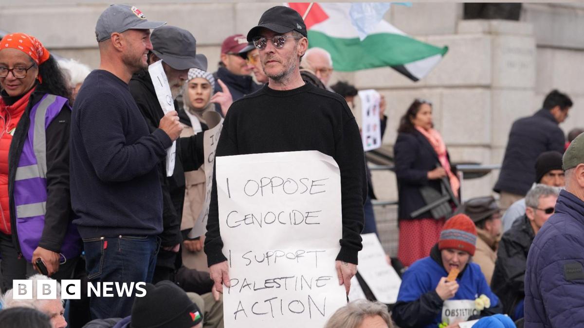 Massive Attack frontman Robert Del Naja standing at a protest at Tralfalgar Square holding a sign that reads: "I oppose genocide I support Palestine Action." He is wearing a black baseball cap, sunglasses and a black jumper
