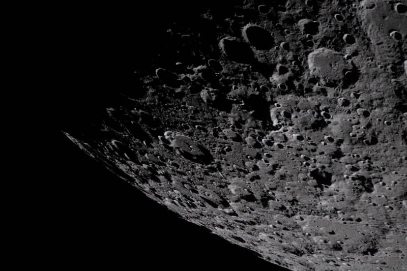 A close-up view of the moon's heavily cratered surface with deep shadows, showing rugged terrain and numerous impact craters against the darkness of space.