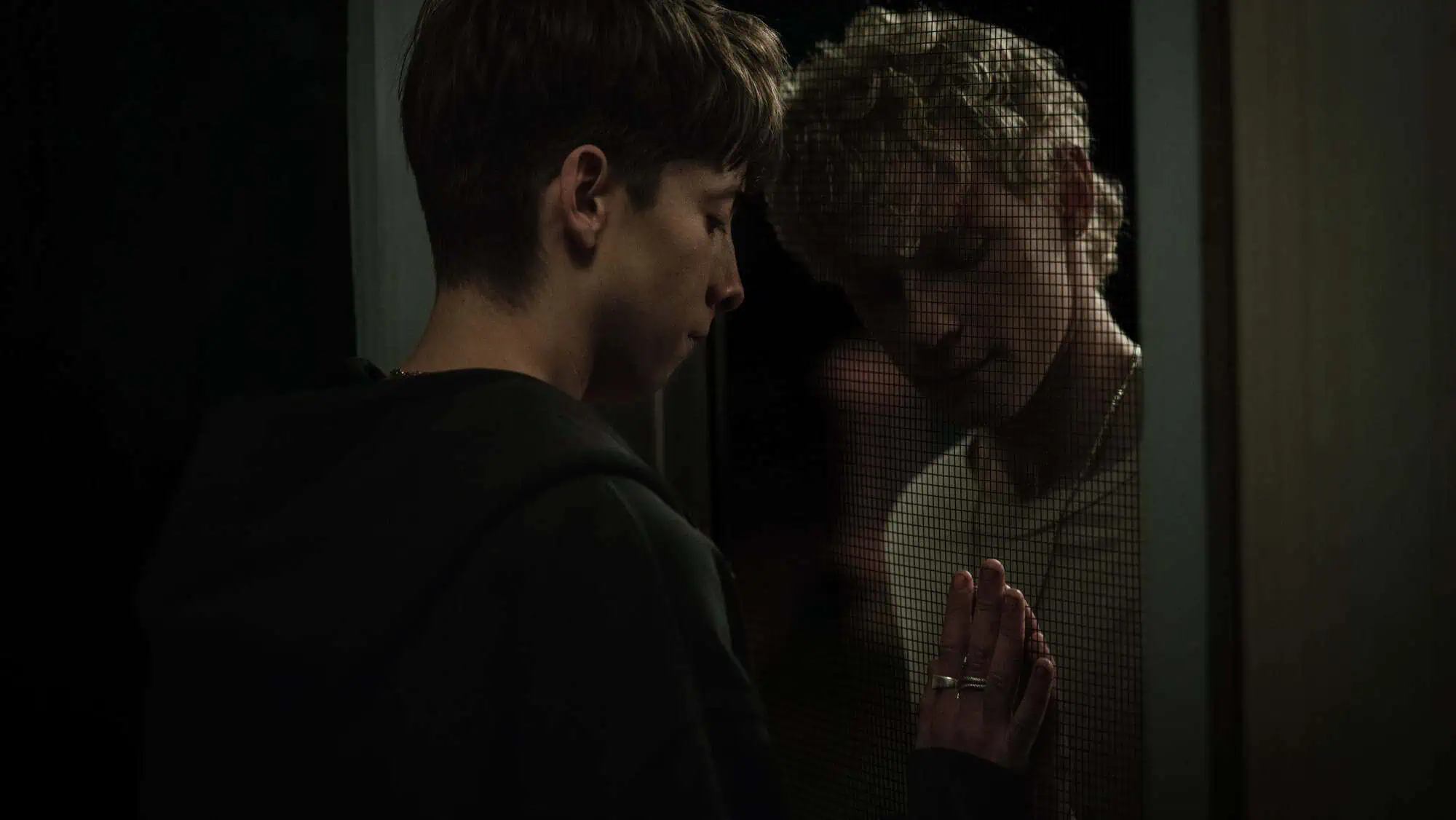 Two young people stand on opposite sides of a mesh screen door, touching hands through the barrier and gazing at each other with somber expressions in dim lighting.