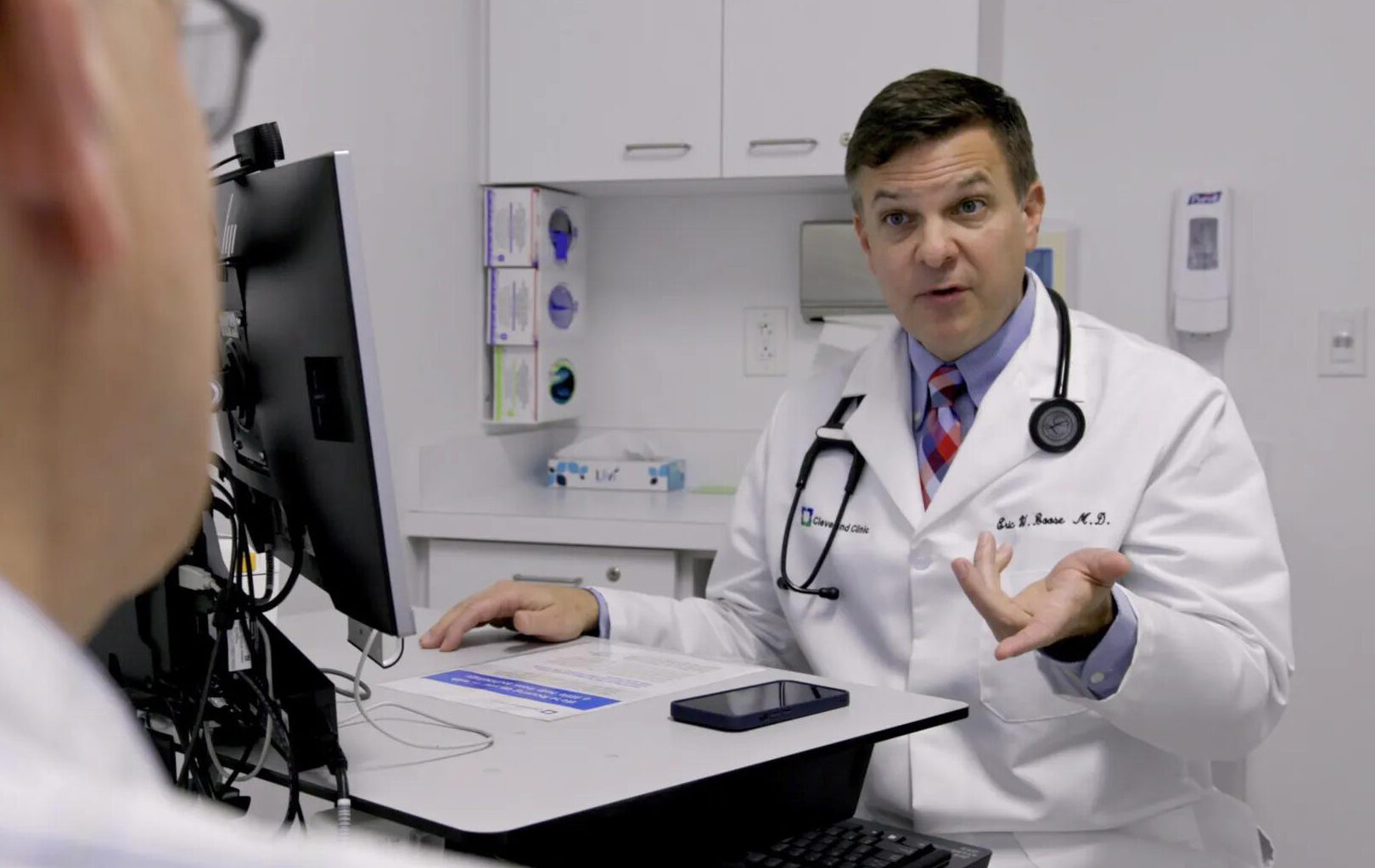 A male doctor in a white coak speaks to someone out of frame.