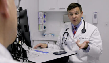 A male doctor in a white coak speaks to someone out of frame.