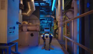 A four-legged Boston Dynamics robot called Spot walks down the hallway of an industrial facility. A human worker wearing a bright yellow safety vest is starting to cross the hallway behind the robot.