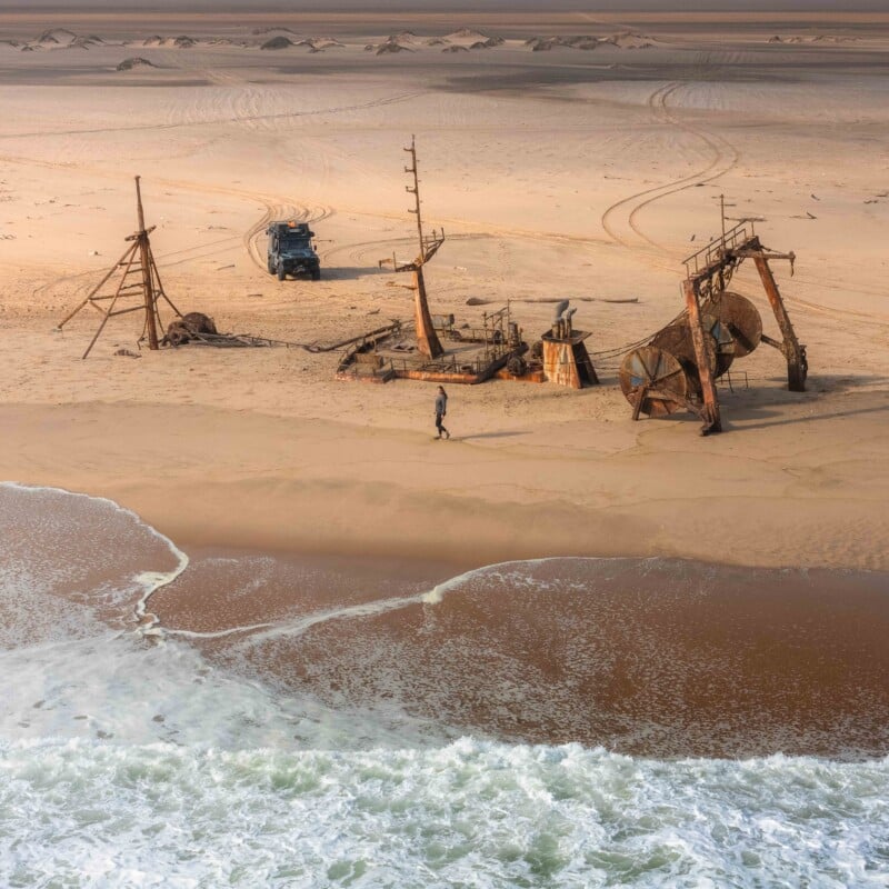 A person walks near the rusted remains of a shipwreck on a sandy beach, with ocean waves in the foreground and a vehicle parked in the background on the dunes.