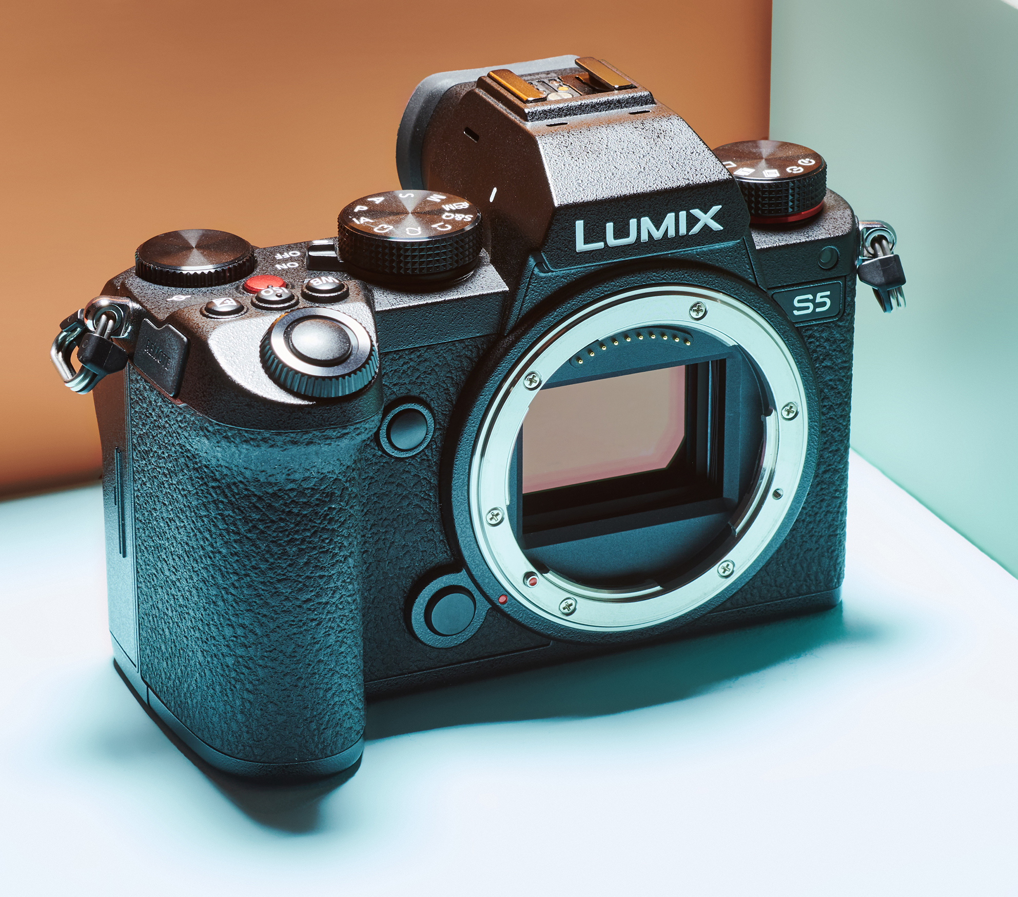 A black Lumix S5 camera body is displayed without a lens. It sits on a light surface against a soft orange and white backdrop