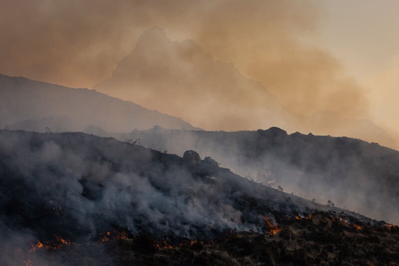 A smoky wildfire burns through dry grass on a mountainside, with flames visible in the foreground and a hazy mountain peak looming in the background under a sky filled with smoke.