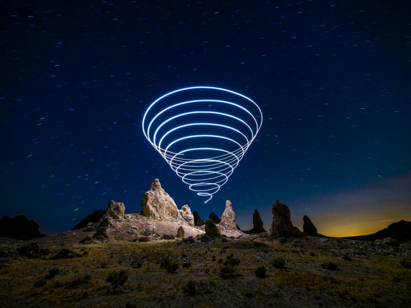 Rock formations under a starry night sky, with spiral-shaped light trails hovering above, creating a glowing cone effect in the sky. The landscape appears arid with sparse vegetation.