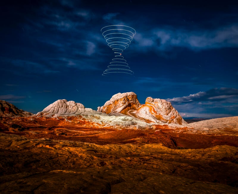 Rocky, desert landscape under a blue, cloudy sky at night. In the sky above the rock formations, a spiral pattern of white lights forms a double-cone shape, resembling a vortex or hourglass.