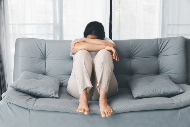 A depressed woman sits curled up on a gray sofa by a window, hiding her face in her arms. This poignant image captures themes of mental health, loneliness, and emotional distress in a bright room.