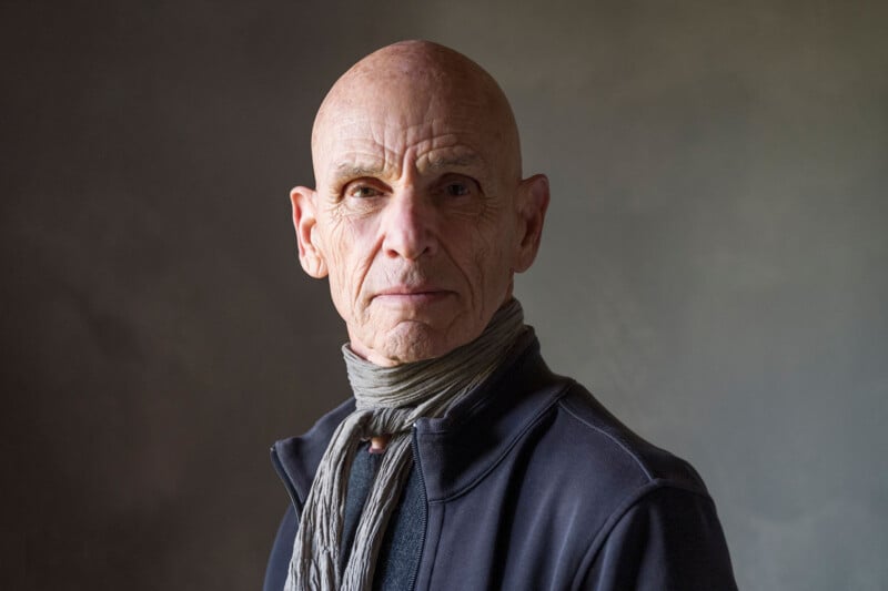 An older, bald man with a serious expression, wearing a dark jacket and a light gray scarf, stands against a plain, softly lit background.