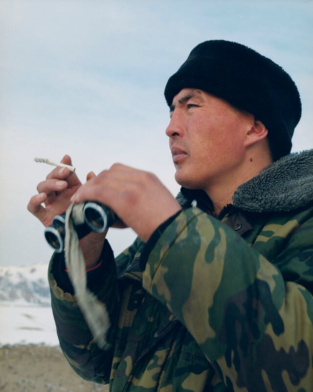 A person in a camouflage jacket and fur hat holds binoculars in one hand and a cigarette in the other, looking into the distance with a snowy mountain landscape in the background.