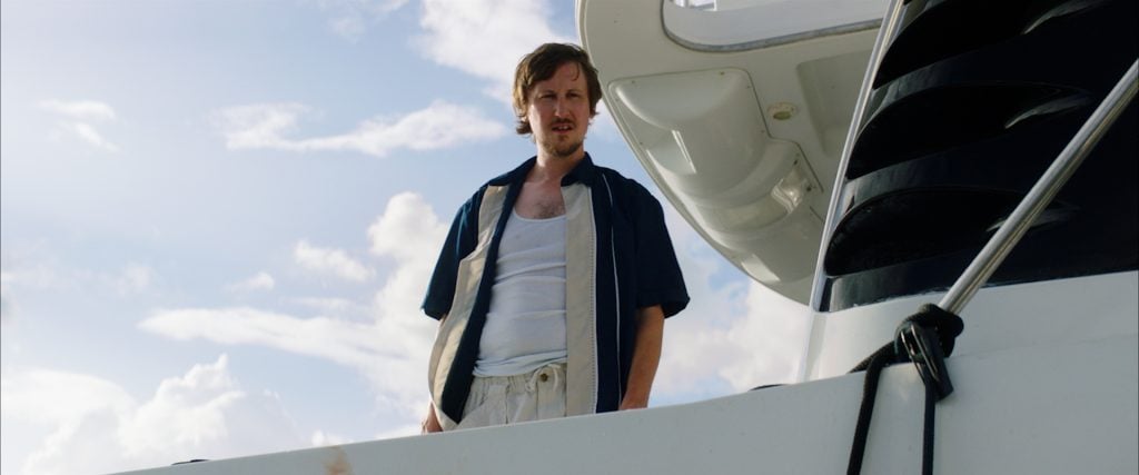 A man in casual clothing stands on the deck of a white boat, looking down with a serious expression against a cloudy sky.