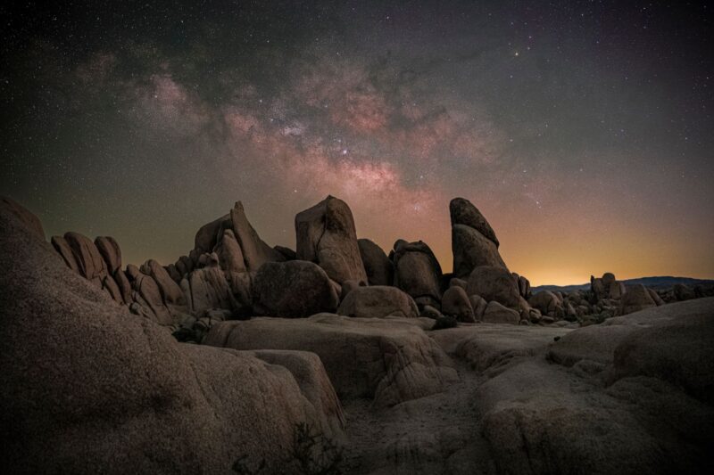 Tall, protruding rock formation with the glowing, billowing Milky Way above, and a glow on the horizon.