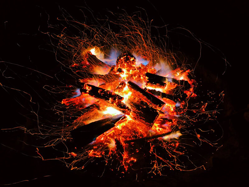 A campfire burns brightly at night, with orange flames, glowing embers, and flying sparks surrounding the stacked logs against a dark background.