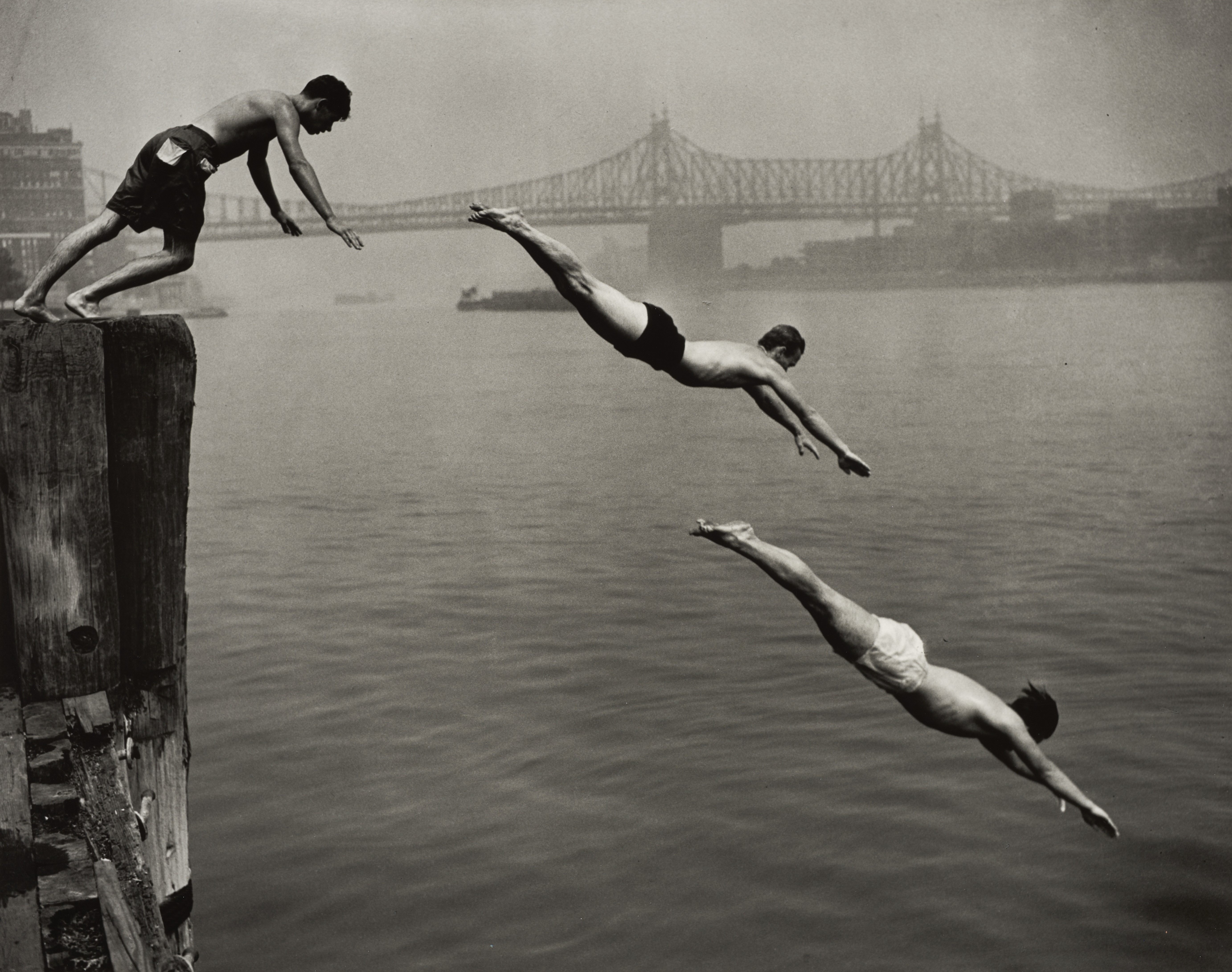 Three young men dive from a wooden pier into the East River, their bodies suspended in mid-air in a descending arc, with the Queensboro Bridge looming through a hazy sky behind them.