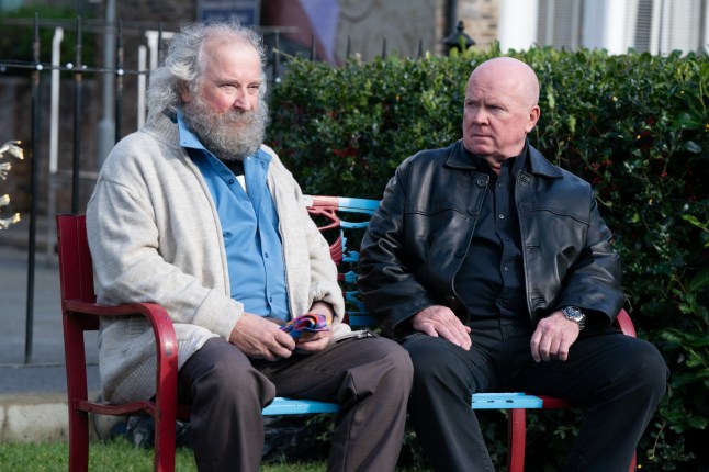Nigel with a full beard sitting next to Phil on a bench in EastEnders