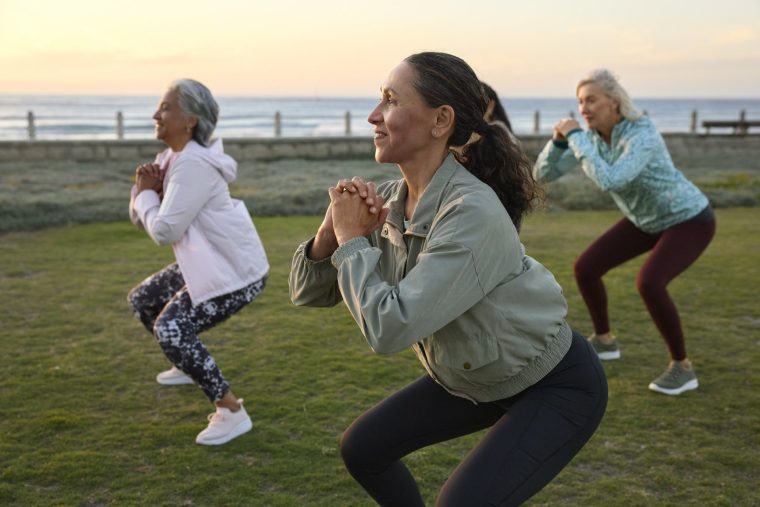 Smiling woman doing exercise with friends in garden. Active female is squatting during morning routine. She is wearing sports clothing.