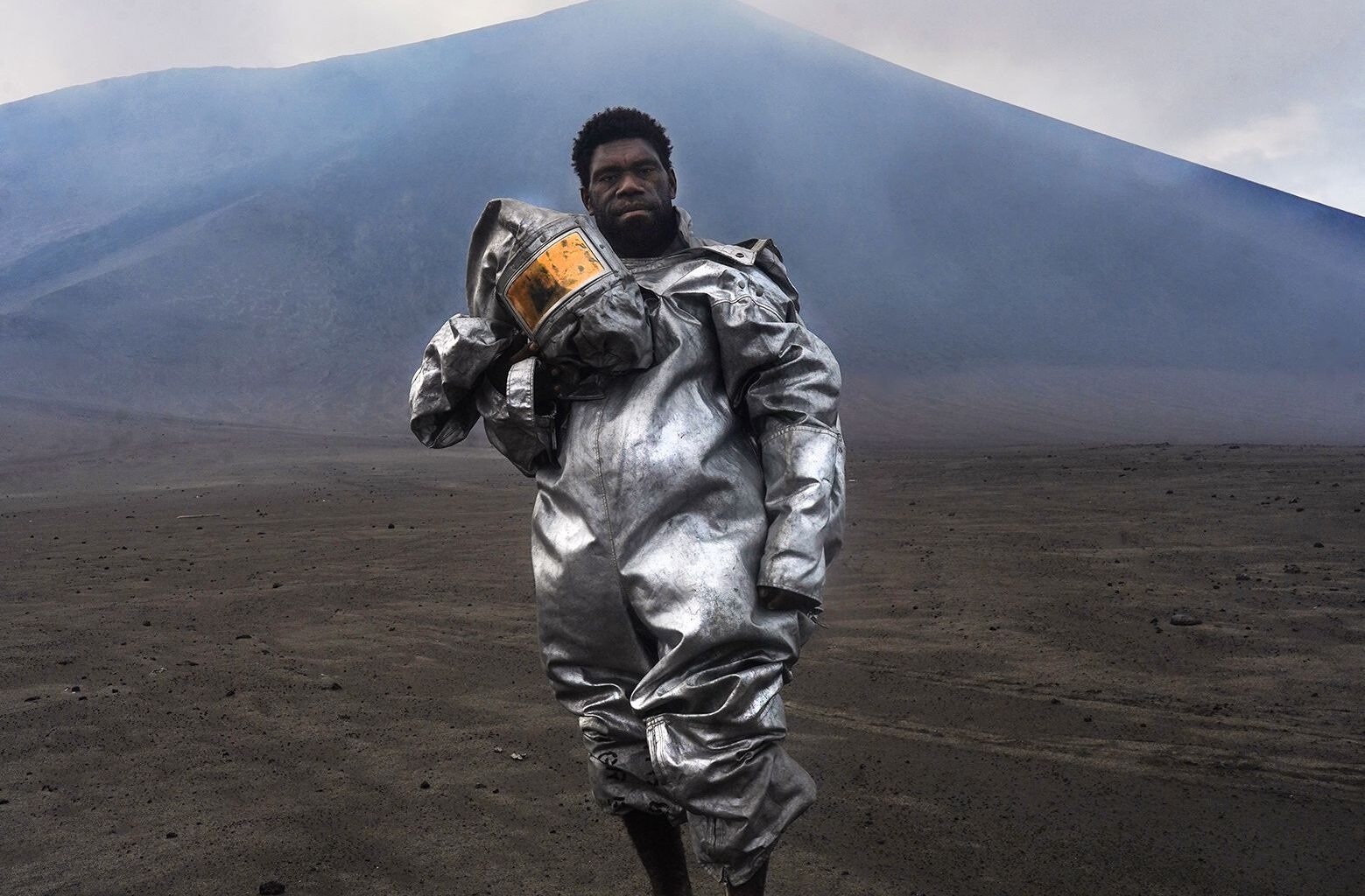 Phillip, a self-taught volcanologist, stands barefoot atop a volcanic rock bomb. Wearing a lava-protection suit, the volcano smoulders behind him, sending a plume of gas and sulphur into the sky