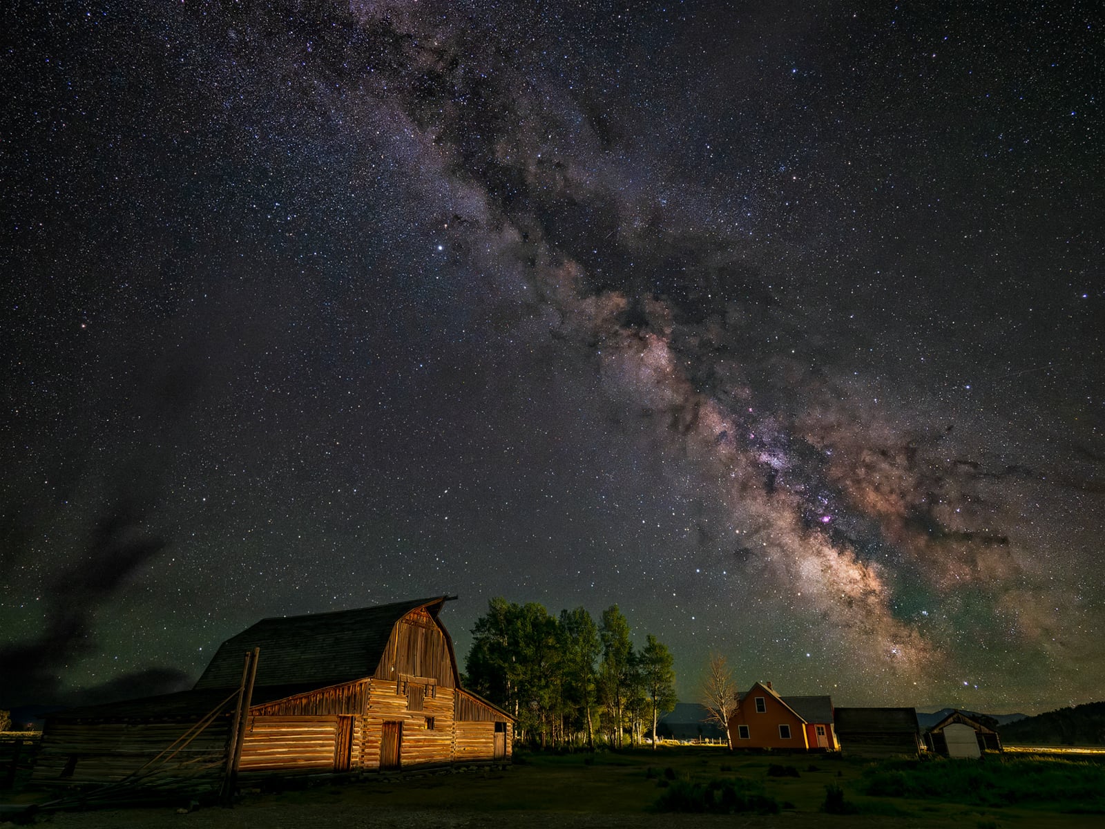 A star-filled night sky with the Milky Way galaxy bright overhead, above a rustic wooden barn and several small buildings surrounded by trees in a rural countryside setting.