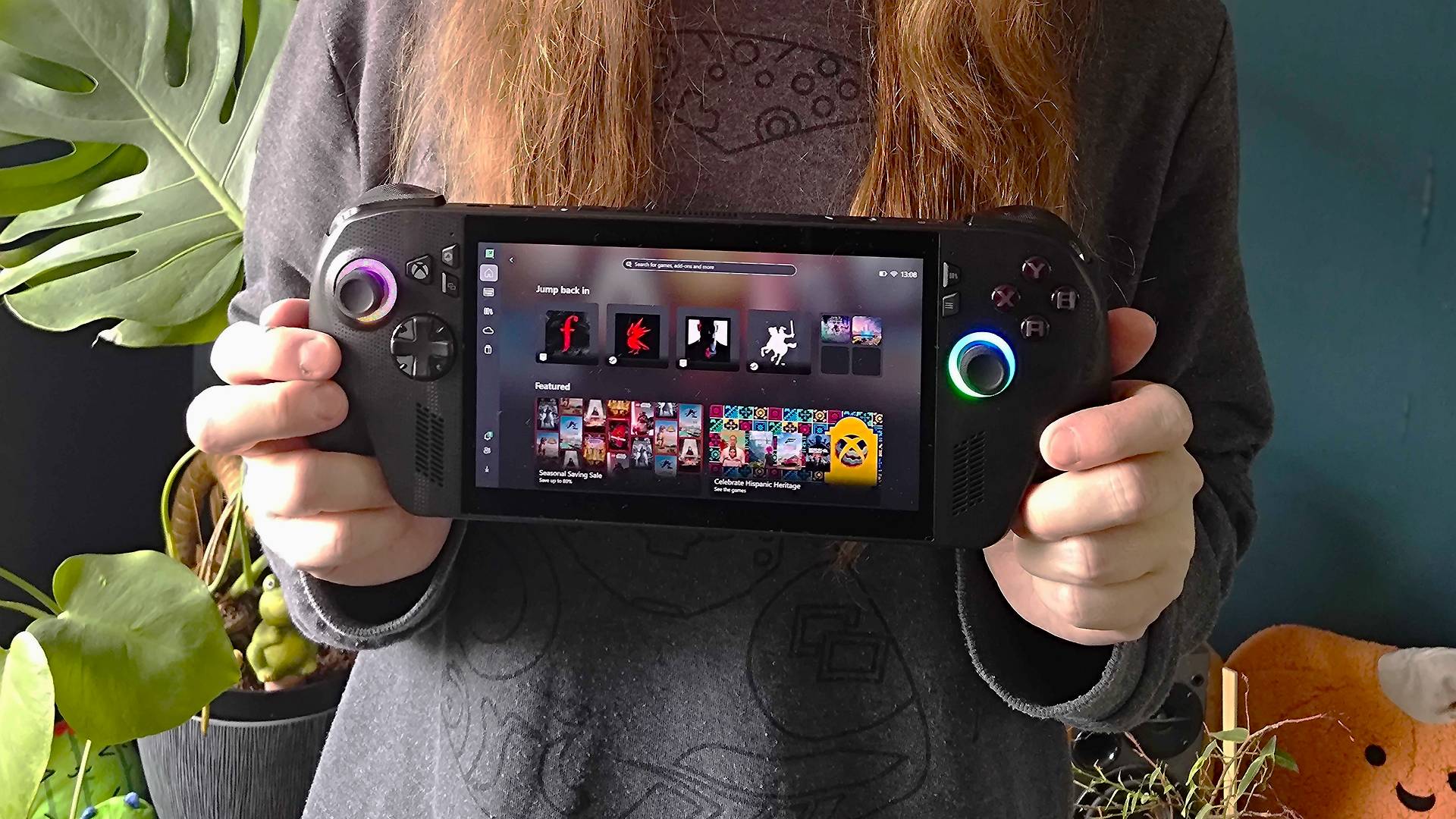 Hands holding ROG Xbox Ally X handheld with plants and green wall in backdrop.