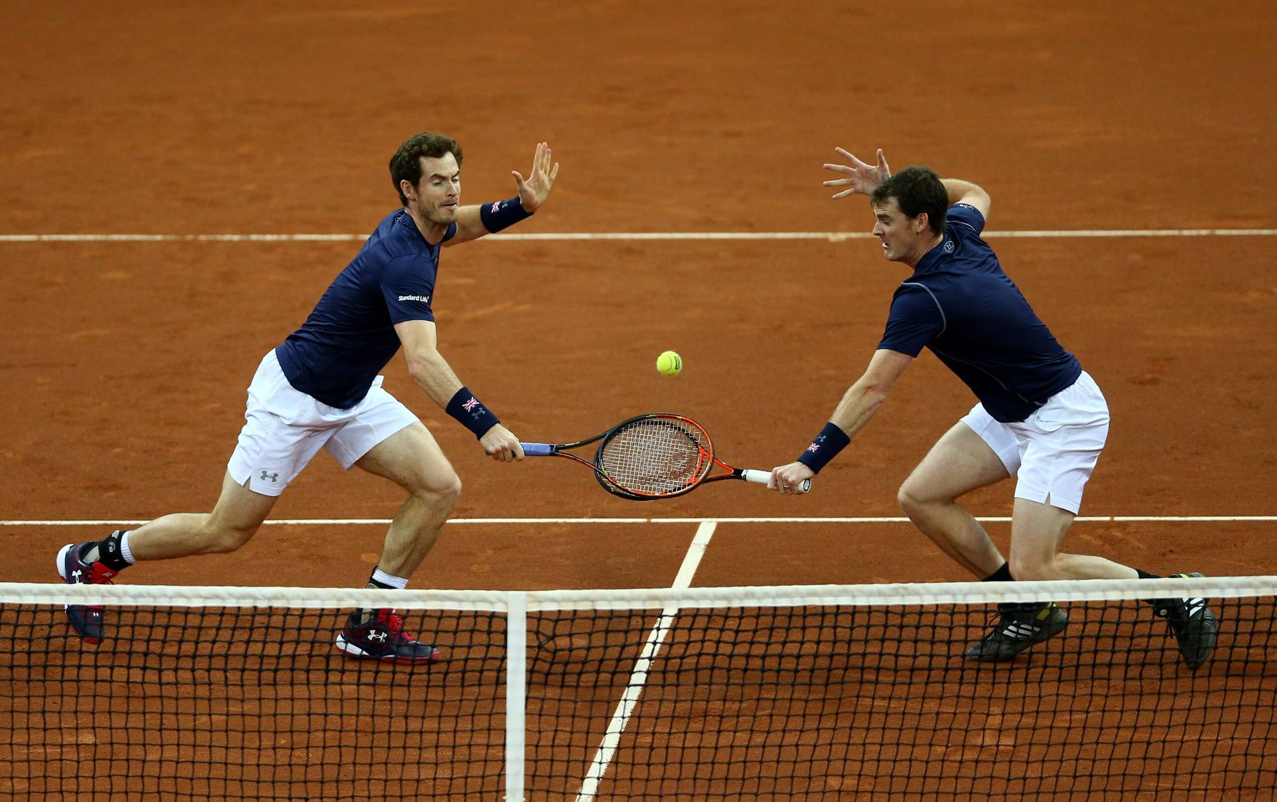 Jamie Murray (right) and Andy Murray (left) both go for a backhand volley down the center of a clay court during a men's doubles match.