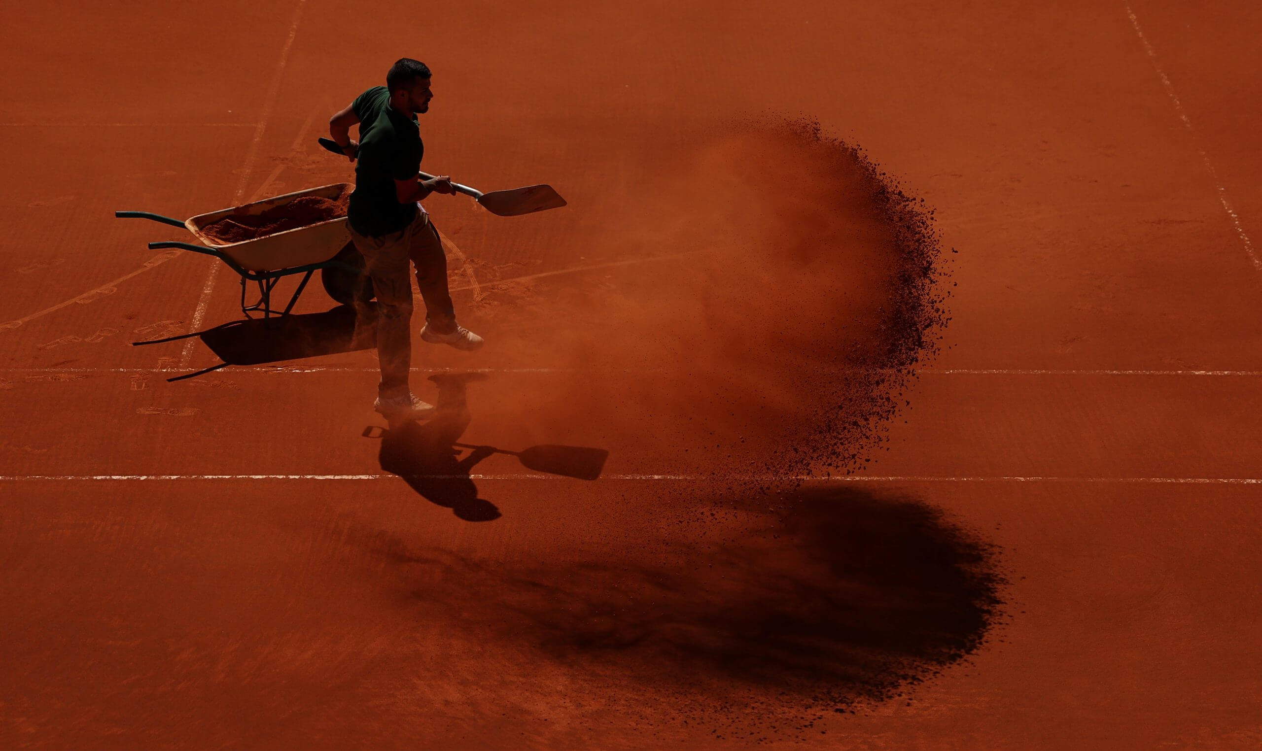 A man scoops clay onto a tennis court with a wheelbarrow to his left full of red brick dust.