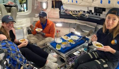 three astronauts wearing blue baseball hats sample foods inside a spacecraft simulator