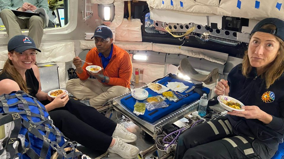 three astronauts wearing blue baseball hats sample foods inside a spacecraft simulator