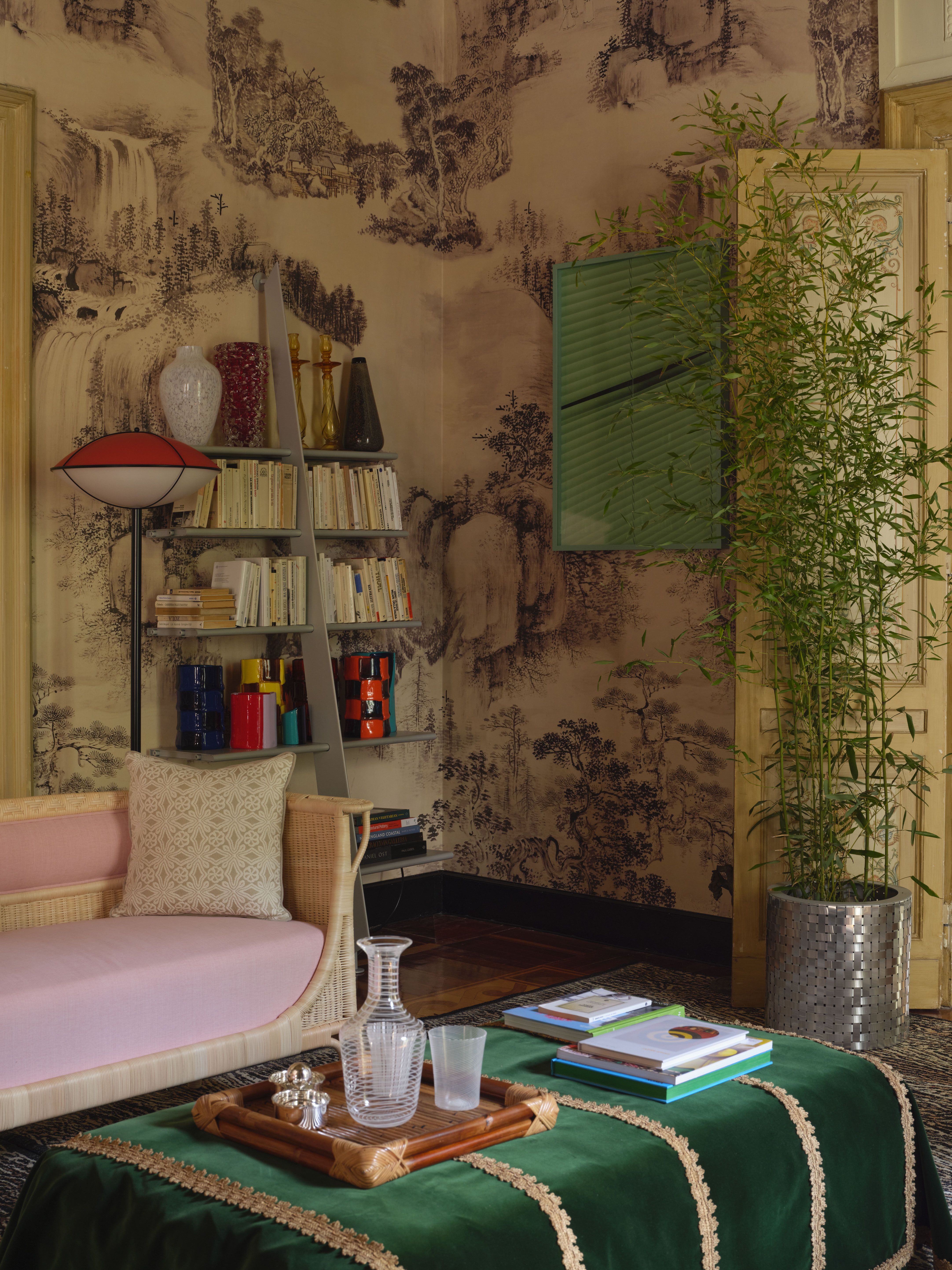 a room with neutral patterned wallpaper, a bookshelf, pink sofa, green stool, and mint artwork. 