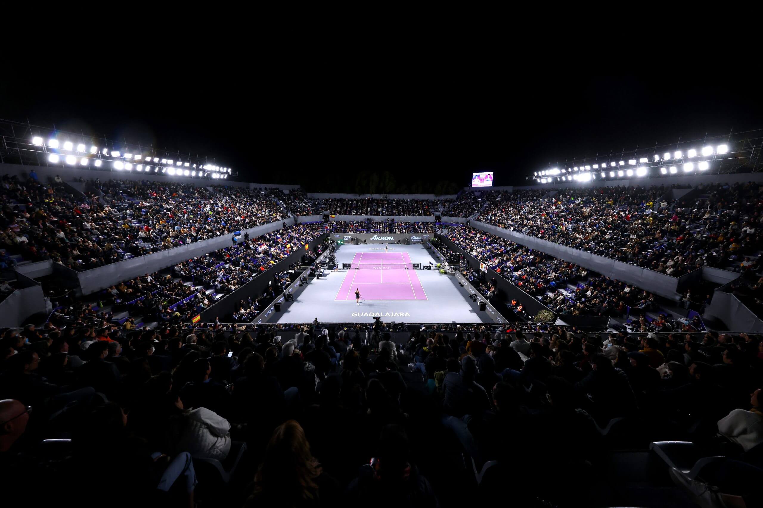 A full tennis stadium at night with floodlights, with a purple hard court.