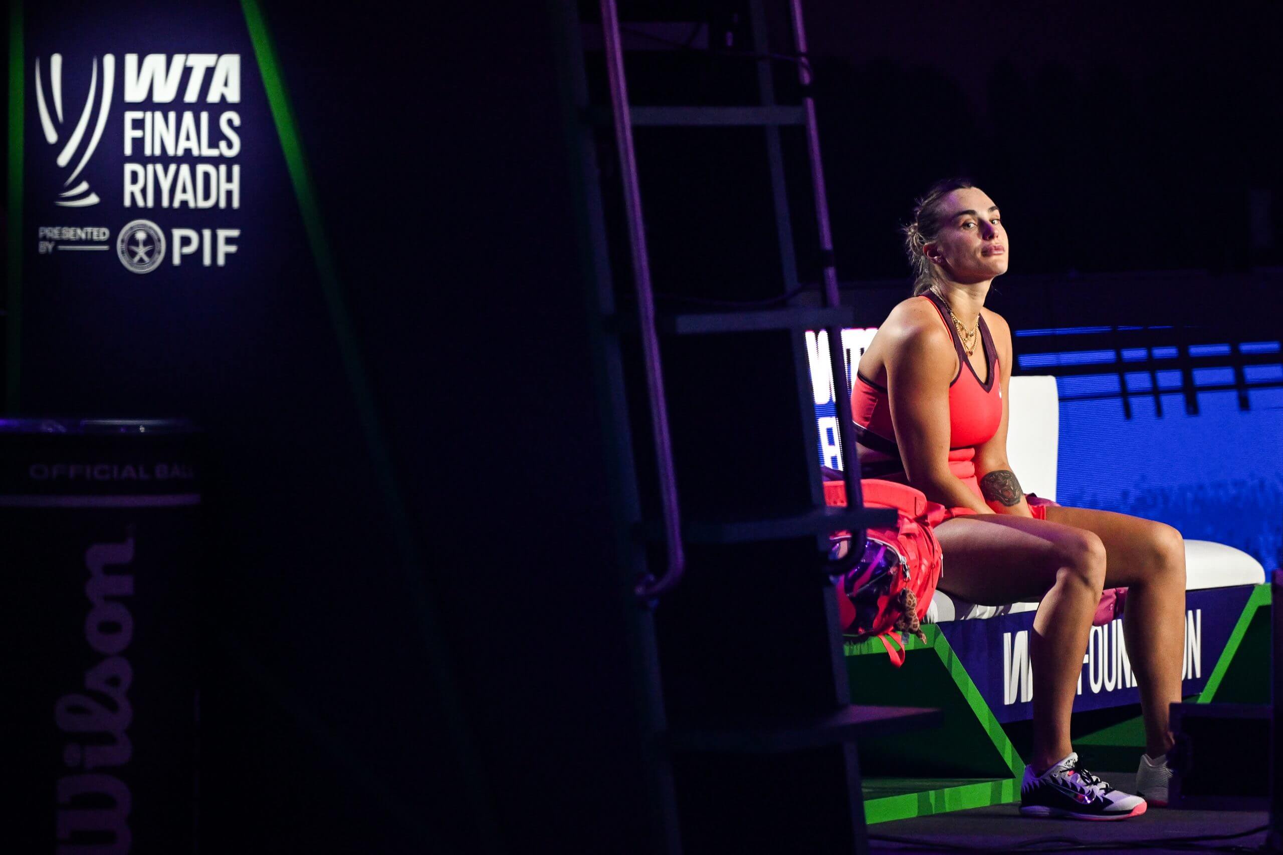 Aryna Sabalenka sits in purple light to the right of a white logo, displaying the WTA Finals Riyadh with the PIF logo.