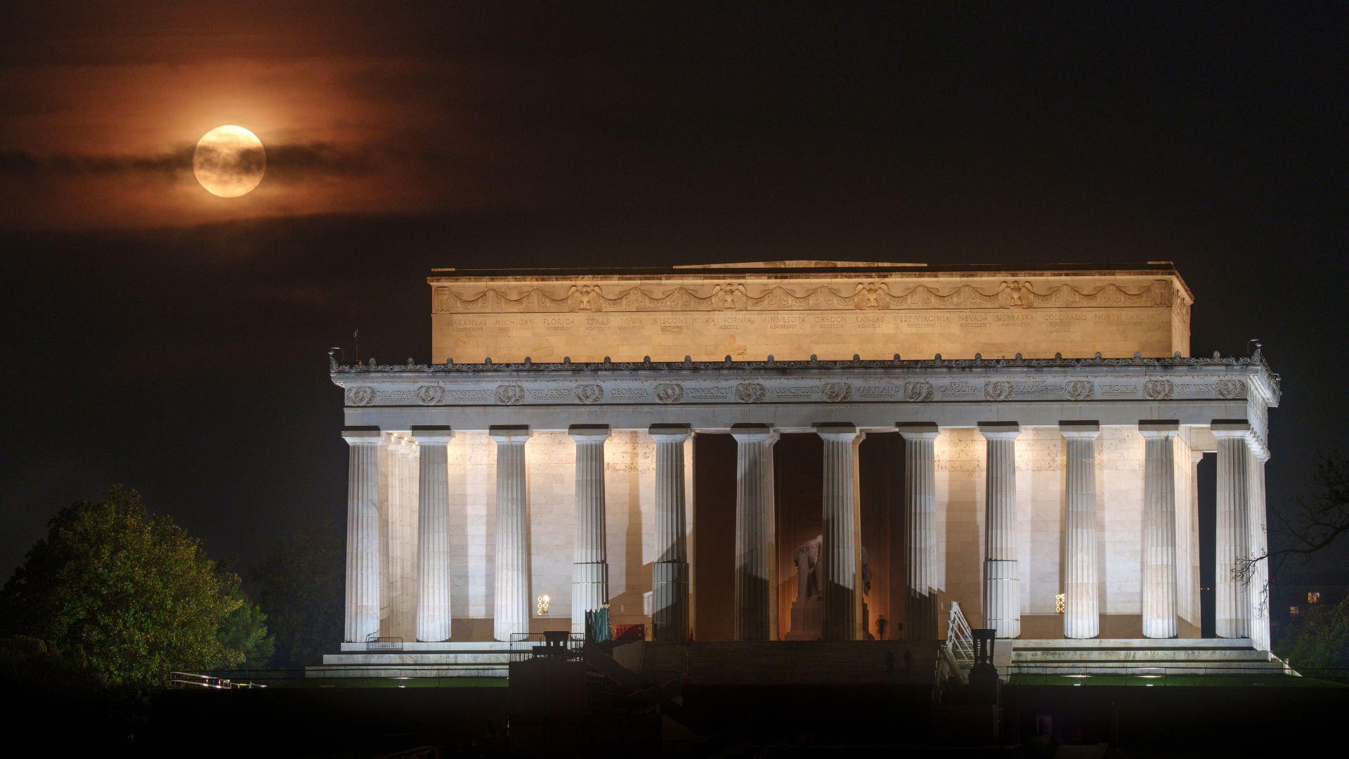 A yellow full moon is pictured in a cloudy sky at night, shining above the columned Lincoln Memorial in Washington D.C.
