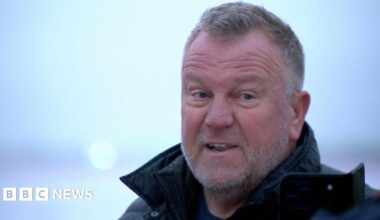 The photo shows four astronauts from NASA’s Artemis II Moon mission standing in a busy indoor sports arena, wearing bright blue flight jackets with mission patches and logos. They are in the foreground, smiling and cheering, with one woman in the centre clasping her hands and two of the men raising their fists in the air in celebration. The crowd behind them is slightly out of focus but fills the stands, suggesting a major sporting event. The lighting is bright and even, and the mood is joyful, energetic and triumphant.