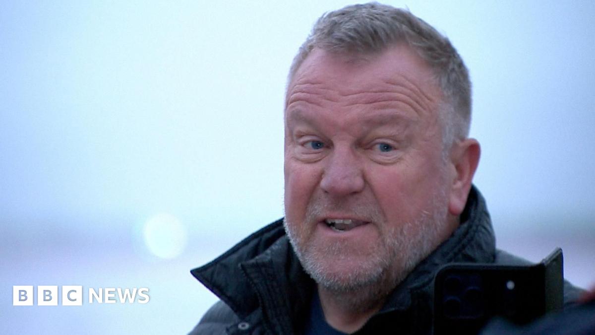 The photo shows four astronauts from NASA’s Artemis II Moon mission standing in a busy indoor sports arena, wearing bright blue flight jackets with mission patches and logos. They are in the foreground, smiling and cheering, with one woman in the centre clasping her hands and two of the men raising their fists in the air in celebration. The crowd behind them is slightly out of focus but fills the stands, suggesting a major sporting event. The lighting is bright and even, and the mood is joyful, energetic and triumphant.