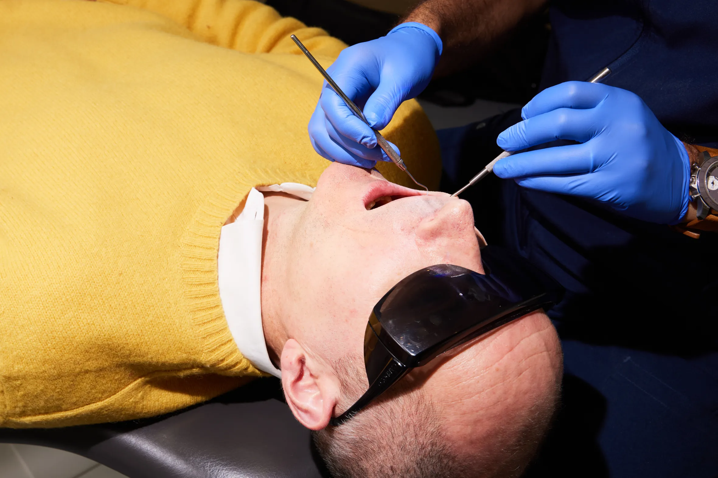 A person in a yellow sweater with protective glasses open their mouth while a dentist in blue gloves examines it with dental tools.
