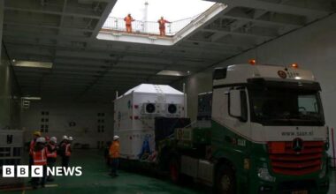 A large, white container on the back of a lorry. It is inside a ship's cargo hold and is surrounded by workers in high-visibility clothing.