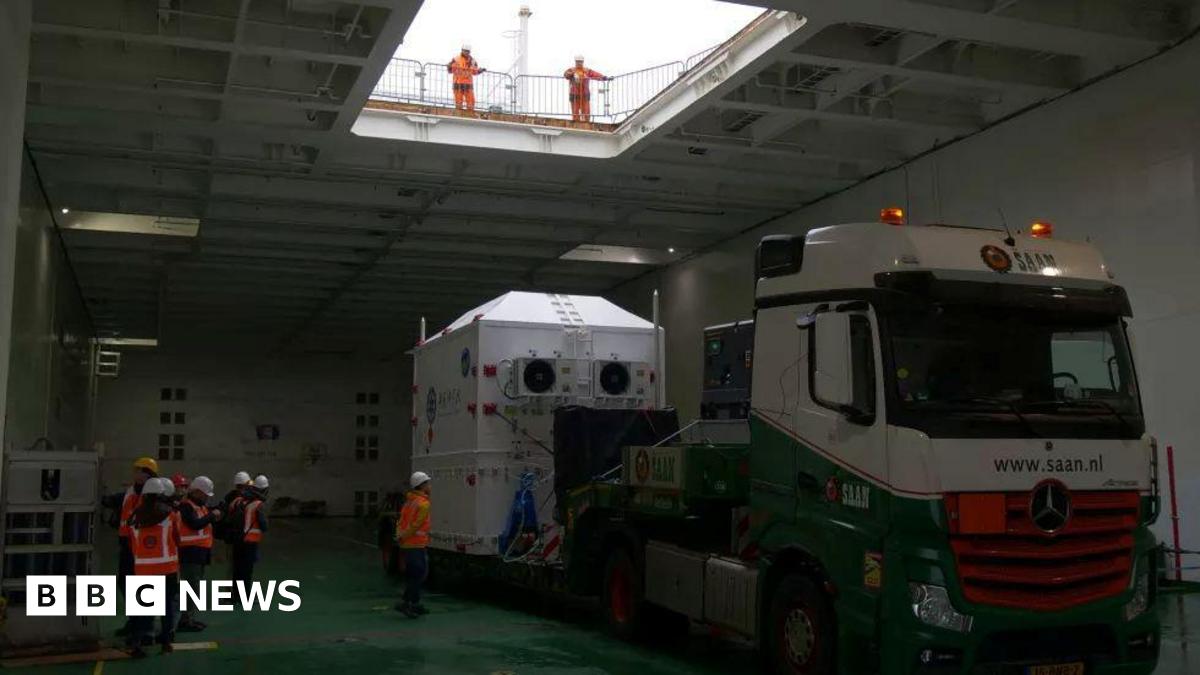 A large, white container on the back of a lorry. It is inside a ship's cargo hold and is surrounded by workers in high-visibility clothing.